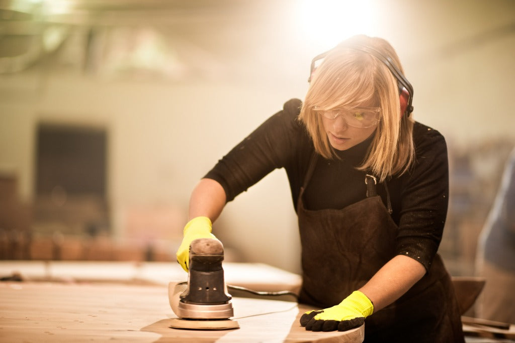 Craftperson with protective equipment working with power sander in workshop making furniture parts.