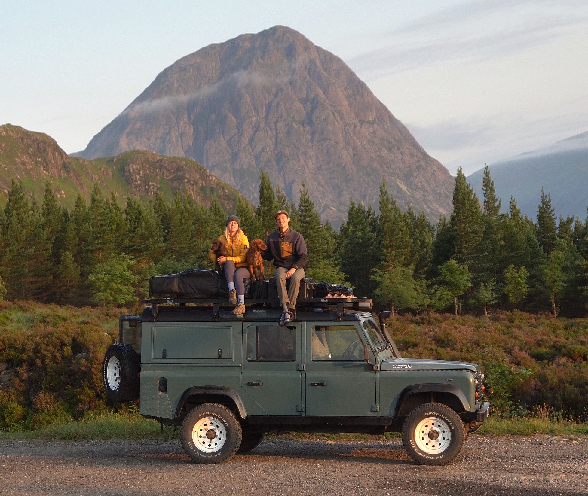 A woman, man, and their dog sit on top of their parked car while on a camping trip. 