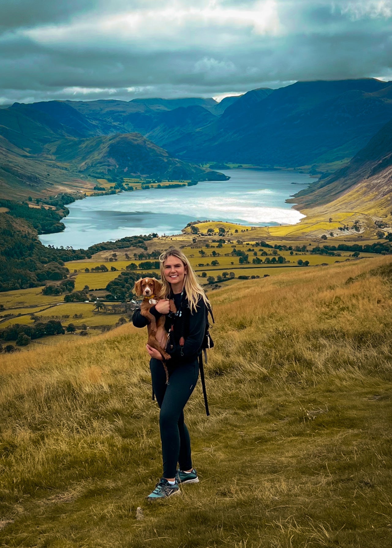 Woman holding a spaniel puppy on a grassy hillside above a lake.
