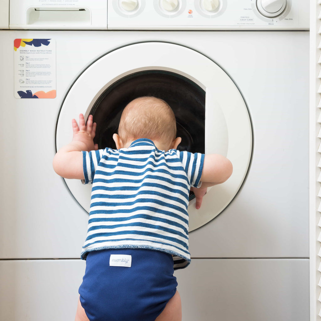 Little boy wearing blue striped shirt and blue Esembly Cloth Diaper looking into washing machine