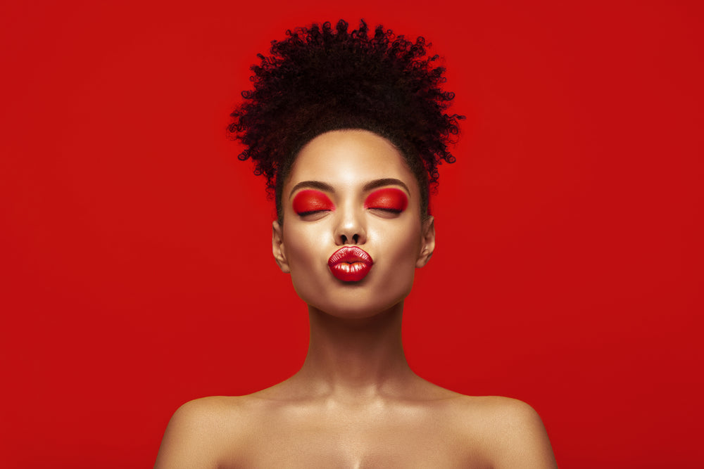 beautiful african american woman on red background with naturally coily hair pulled up into pineapple style  with artistic red eyeshadow blowing a kiss