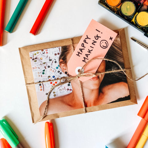 A package wrapped in brown paper and twine with a colourful photo of a woman with lots of earrings around her hair. The package is surrounded with art materials such as textas and paints. It's laying on a white background