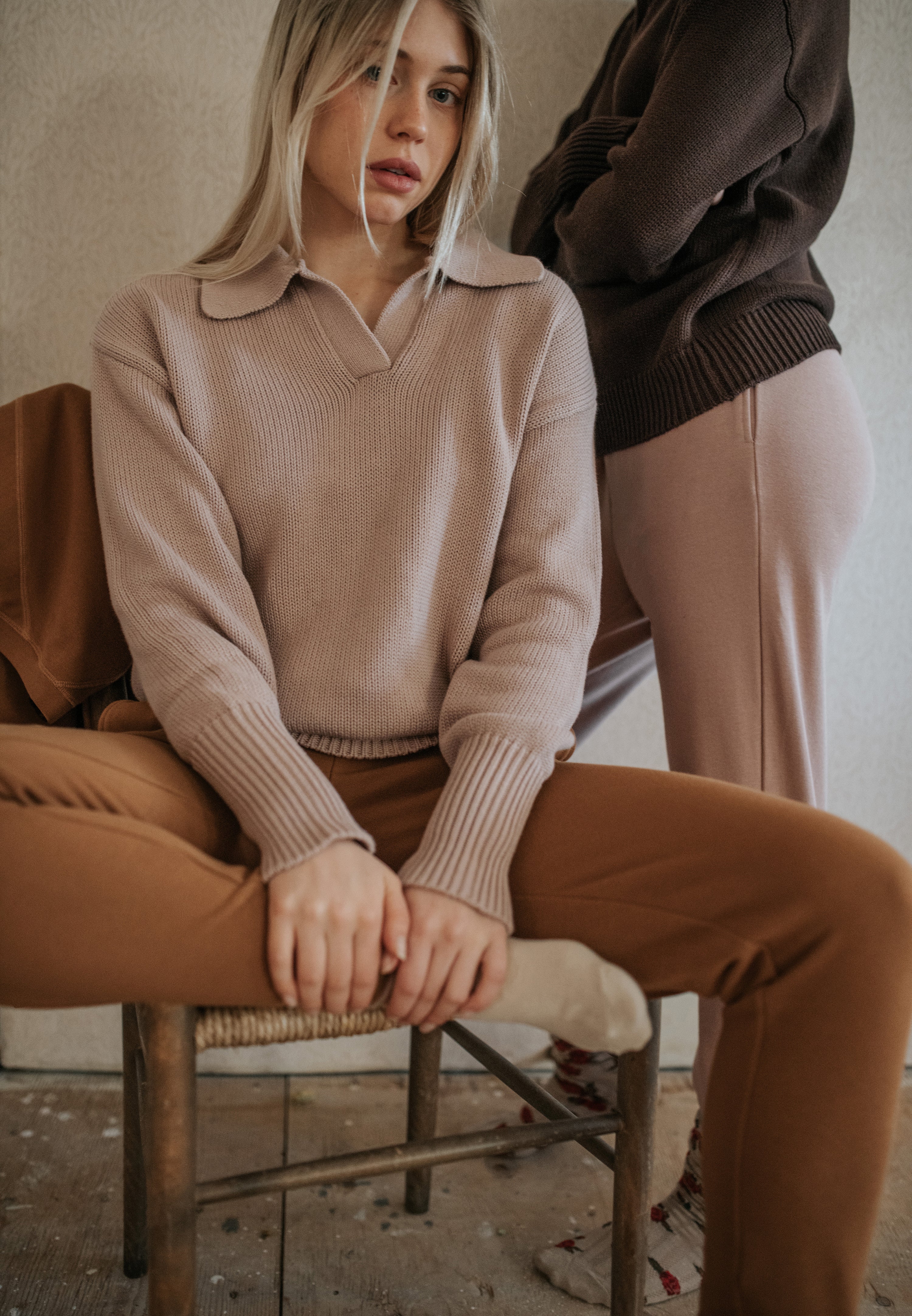 one woman sits on a chair in knit lounge pants and a knit collared sweater.  Another women stands behind in coordinating colours of the same outfit. 