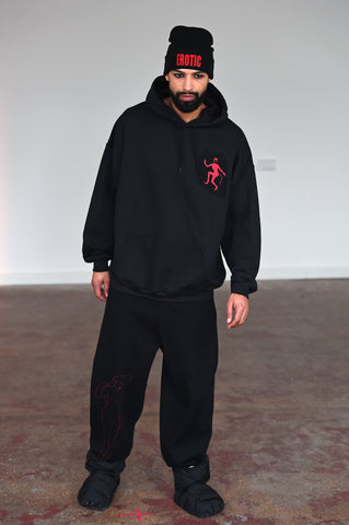 LULA LAORA AW21 Getty Images, menswear model wears a black beanie with the words erotic in red. the hoodie is black with a Red Devil lady logo, he also wears black sweatpants or joggers and puffy black shoes. 