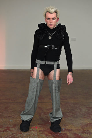 LULA LAORA AW21 Getty Images, menswear model wears a black ruffled harness over a long sleeved black t-shirt and black underwear. He also wears puffy shoes and houndstooth garter trousers. 