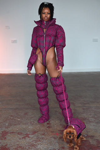 LULA LAORA AW21 Getty Images, womenswear model wears pink fine houndstooth puffy shoes that are thigh high. She also wears a small pink houndstooth bag and a puffer jacket bodysuit with a high leg in pink. She also holds the leash of a dog wearing a matching pink houndstooth jacket. 