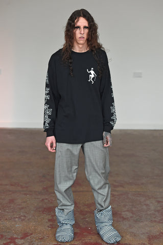 LULA LAORA AW21 Getty Images, menswear model wears houndstooth trousers with matching puffer shoes. Also wears a black long sleeved t-shirt with ivy illustrations on the sleeves and a white evil lady logo. 