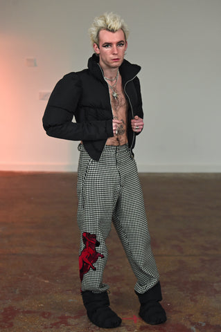 LULA LAORA AW21 Getty Images, menswear model wears houndstooth trousers with red embroidered ram on the leg. he wears puffy black does. he also wears a short black puffer jacket that he holds open. 