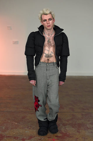 LULA LAORA AW21 Getty Images, menswear model wears houndstooth trousers with red embroidered ram on the leg. he wears puffy black does. he also wears a short black puffer jacket that he holds open. 