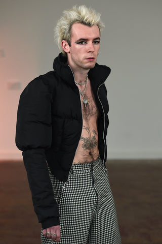 LULA LAORA AW21 Getty Images, menswear model wears houndstooth trousers with red embroidered ram on the leg. he wears puffy black does. he also wears a short black puffer jacket that he holds open. LULA LAORA AW21 Getty Images, menswear model wears houndstooth trousers. he wears puffy black does. he also wears a short black puffer jacket.