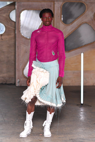 Lula Laora runway Getty pink eyebrows, menswear, mesh long sleeved fuchsia top with a high neck and embroidered beetles on it. white high top sneakers with lace up closing. Aqua light blue skirt with white ruffles. 