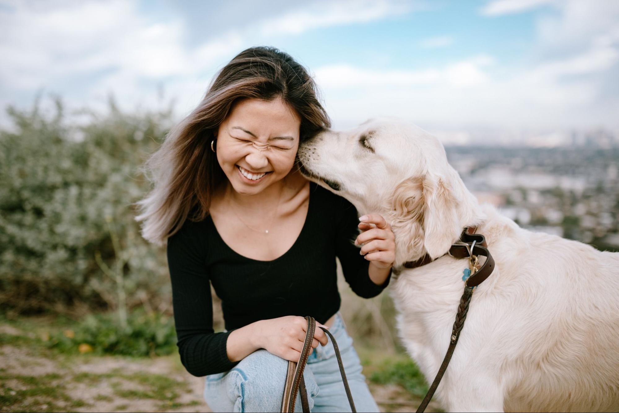 woman smiling while dog licks her face in a field