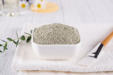A bowl of fine, gray bentonite clay powder displayed on a white towel next to a brush with a wooden handle and green eucalyptus leaves, suggesting use for a spa or skincare routine.