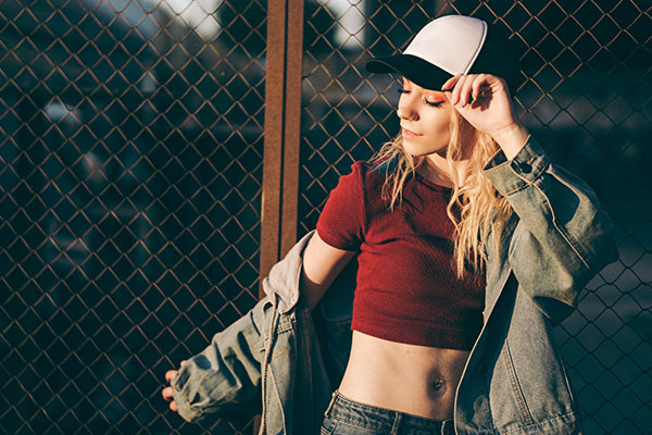 Young attractive woman in jeans jacket, shorts, red top and trucker hat posing over metal fence