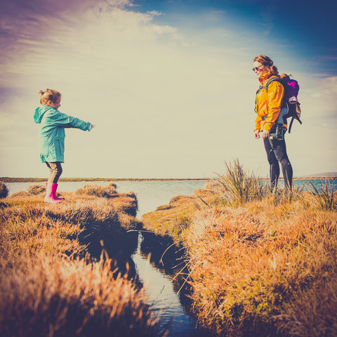 A parent and her child stand slightly apart on two cliffs. The parent is allowing the child to take some risks as advocated by the Slow Parenting movement 
