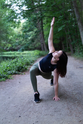 woman wearing sustainable black crop-top and olive leggings doing a yoga pose on a nature park track