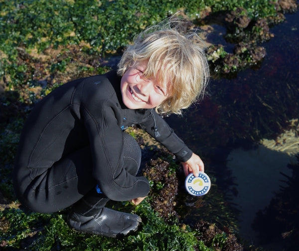 Things to look for in kids sunscreen - a young boy wearing a wetsuit smiles at the camera while crouching beside a rock pool holding a tin of SunButter sunscreen