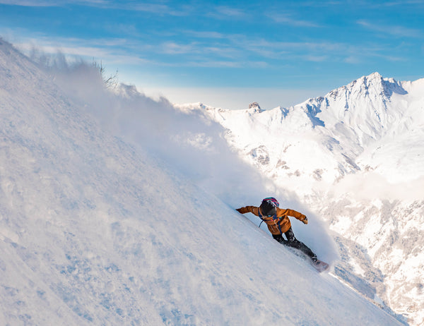 Snowboarder Michaela Davis-Meehan. A figure dressed in an orange snow jacket carves some snow on the side of a mountain with a blue sky in the background.  