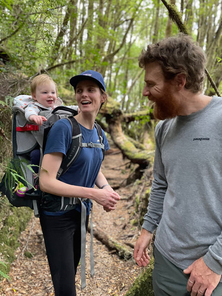 Beau and his partner walk through the bush with their infant daughter, May, in a carrier on her mother's back. All three are smiling. 