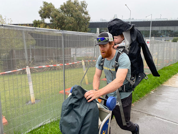 Beau Miles leaves an airport with a trolley and his infant daughter in a carrier on his back 