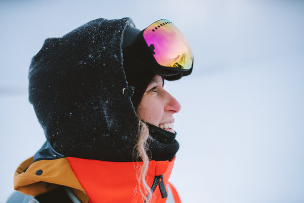 Snowboarder Michaela Davis-Meehan. A side-on close-up image of Michaela wearing red and black snow gear with a hood and snow goggles. She is smiling and there is a wavy piece of hair escaping her hood.  