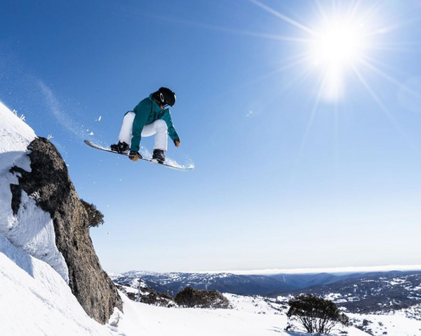 Snowboarder Michaela Davis-Meehan. A snowboarder is airborne after launching off a clifftop in the snow. The sky is blue and the sun shines brightly. 