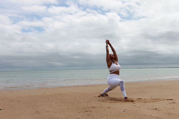 How Ayurveda is useful in modern life. A dark-skinned woman is on a beach wearing white yoga clothes and practicing a yoga pose where she is lunging with her arms above her head.  