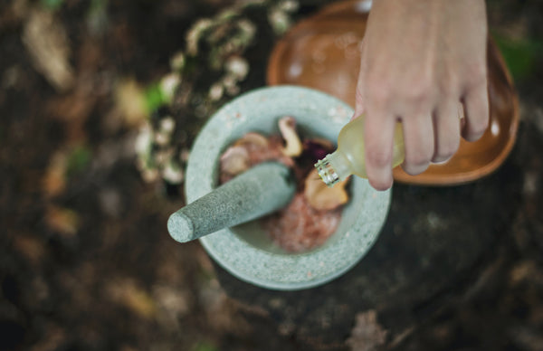 Ayurveda for skincare. A fair-skinned hand pours drops of oil into a mint-coloured mortar and pestle containing other ingredients, which are blurred out. The mortar and pestle is surrounded by greenery.