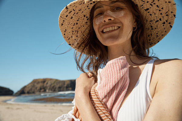 The lowdown on free radicals, antioxidants and your skin. A young woman stands on the beach wearing a straw hat and smiling with zinc across her nose