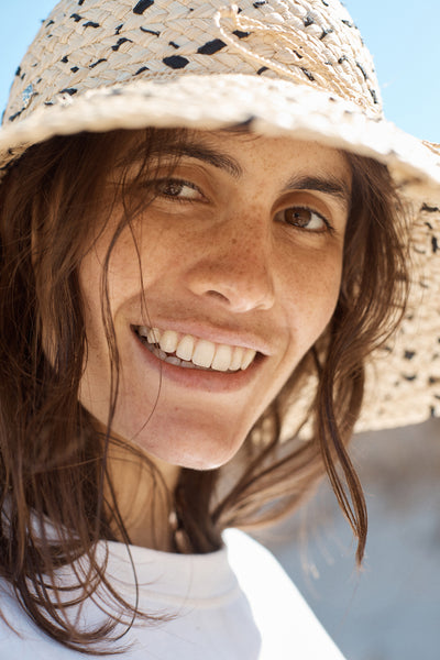 Tinted sunscreen - your questions answered. A young woman with olive skin and brown eyes and hair wear a sun hat and smiles at the camera. 