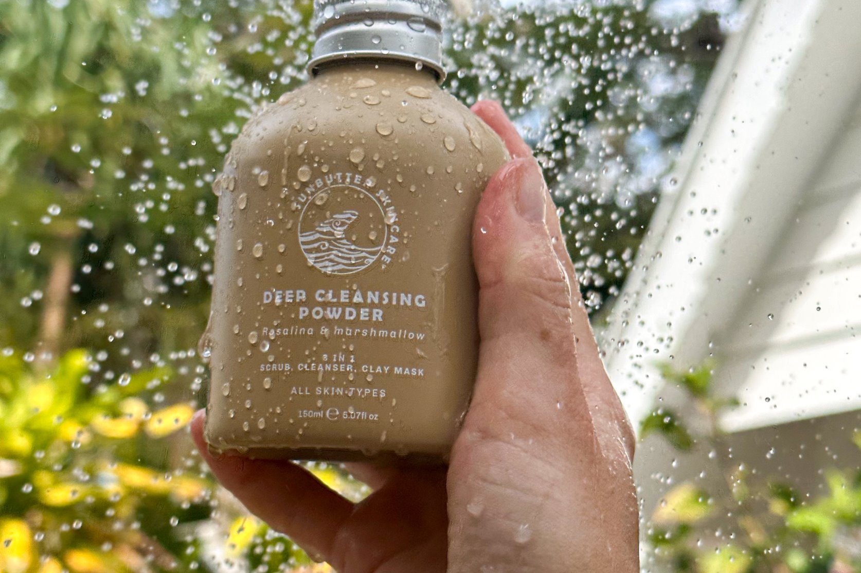 A hand holds a beige coloured bottle that is covered in water droplets against a background of greenery.