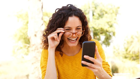 Woman laughing while touching her glasses and using her cell phone.