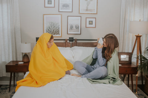 Man wrapped in yellow blanket sitting on bed with woman taking his picture