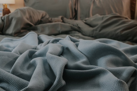 an unmade bed with a blue throw blanket and gray sheets