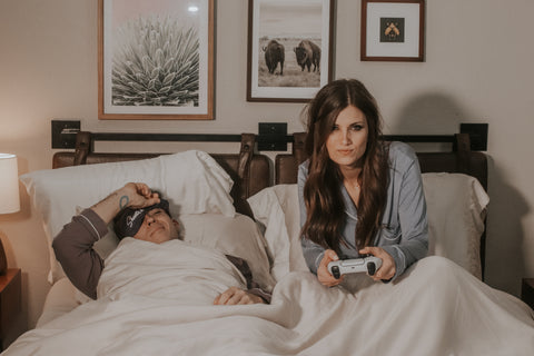 Woman holding video game controller in bed next to man wearing eye mask