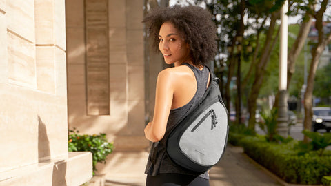 A curly haired woman smiles over her shoulder at the camera. She is carrying Sherpani Anti-Theft sling backpack, the Esprit AT.