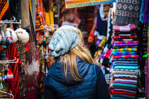 A blonde woman facing away from the camera is looking at the colorful merchandise on display in a market in Peru