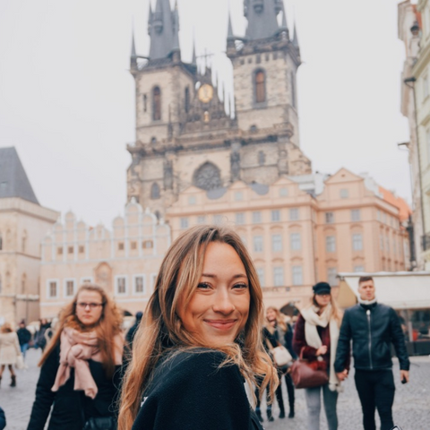 A blonde woman smiles over her shoulder at the camera. She is standing in Prague's Old Town Square, in front of the towering spires of Tyn Church.