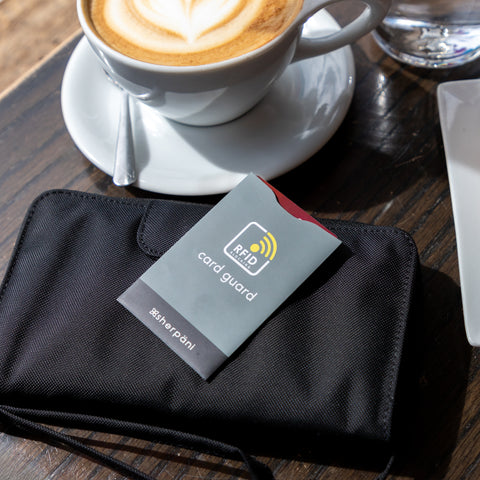 A credit card sits on a table with a coffee and Sherpani RFID blocking wallet, the Tulum. The card is inside Sherpani RFID Credit Card Sleeve to protect the credit card number and prevent cyber theft.