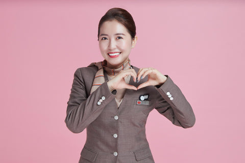 A flight crew member stands in front of a pink background and makes a heart shape with her hands