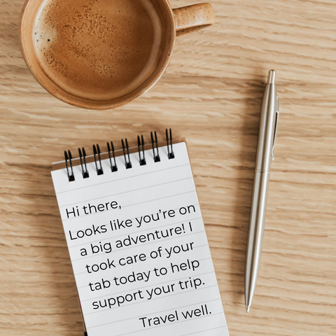 A cup of coffee, notepad and pen sitting on the table. The note reads: Hi there, Looks like you're on a big adventure! I took care of your tab today to help support your trip. Travel well.