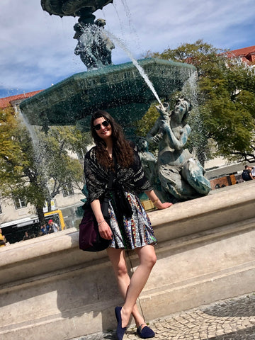 A woman standing in front of a fountain in Lisbon, Portugal. She is wearing a multi-colored dress and Sherpani's Anti-Theft bag the Soleil in Merlot as a crossbody.