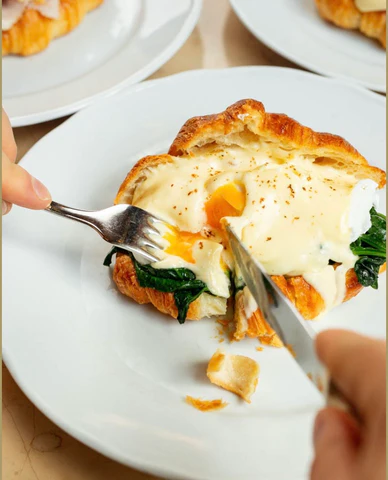 Close up view of hands cutting into a breakfast sandwich with fork and knife.