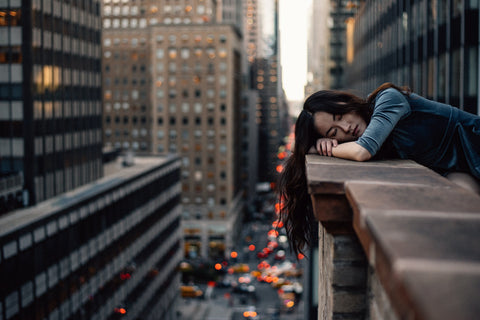 A tired woman leans her head on a ledge with a city view, her eyes are closed