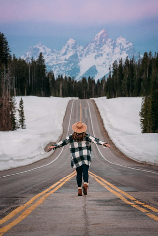 A woman standing in the middle of a road facing a snow-capped mountain with wide open arms