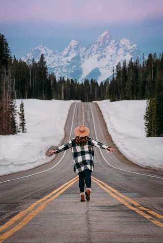 A woman standing in the middle of a road facing a snow-capped mountain with wide open arms