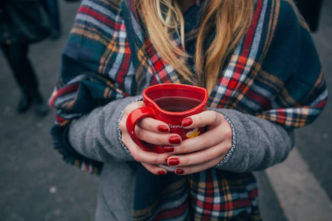 Close up view of a woman's hands clutching a mug. She has blonde hair and is wearing a scarf.