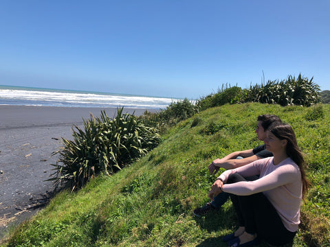 Tess and her bother sit on the grass staring out at the New Zealand ocean and a black sand beach.