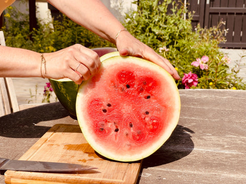 A woman's hands holding a half a watermelon on an outdoor table.