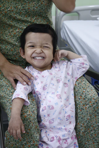 A young Vietnamese girl in a hospital gown. She has a giant grin on her face.